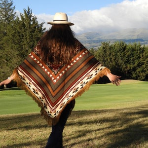 Puede incluir: Una persona lleva un poncho estampado en marrón y beige con flecos. El poncho tiene un diseño geométrico con detalles en rojo, blanco y negro. La persona lleva un sombrero blanco y está de pie en un campo de hierba.