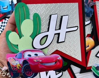 May include: A handmade banner with a red and black border, featuring a silver diamond plate background. The banner has a large white letter "H" and a red race car. Green cactus cutouts add a desert theme.