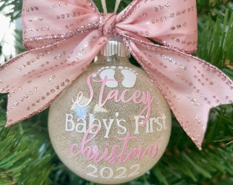 May include: A clear Christmas ornament with glitter inside, featuring the text "Stacey Baby's First Christmas 2022" in pink and white. It is adorned with a pink bow with glitter accents and hangs from a white and gold ribbon.