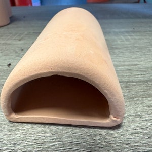 May include: A light pink ceramic hide for reptiles or small animals. The hide has a rounded opening and a smooth, textured surface.