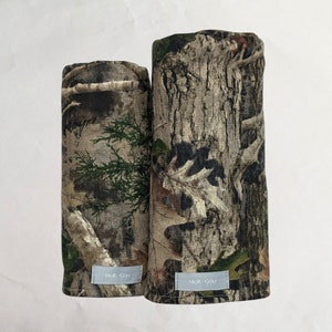 Pode incluir: Duas capas para tacos de golfe com um padrão de camuflagem. As capas são cilíndricas e apresentam uma mistura de tons castanhos, verdes e bege, imitando um ambiente florestal. Cada capa tem uma pequena etiqueta retangular com "Moll Golf".