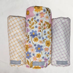 May include: Three fabric swaddles, one with a floral pattern in shades of pink, yellow, and blue, and two with a checkered pattern in yellow and white, and purple and white.