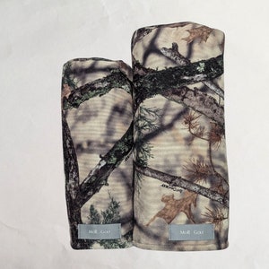 May include: Two camouflage golf headcovers with the text "MOR Golf" on the bottom. The headcovers are in a woodland camouflage pattern with brown, green, and beige colours.