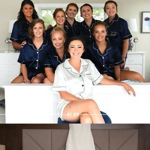 May include: A group of bridesmaids in matching navy blue pajamas with white trim and the word "Bride" embroidered on the front. The bride is wearing a white satin pajama set with black trim. The bridesmaids are sitting on a white bed, and the bride is standing in front of them.