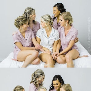 May include: Six women in matching pink pajamas are sitting on a white bed. They are smiling and laughing. Some of the women are holding champagne flutes.