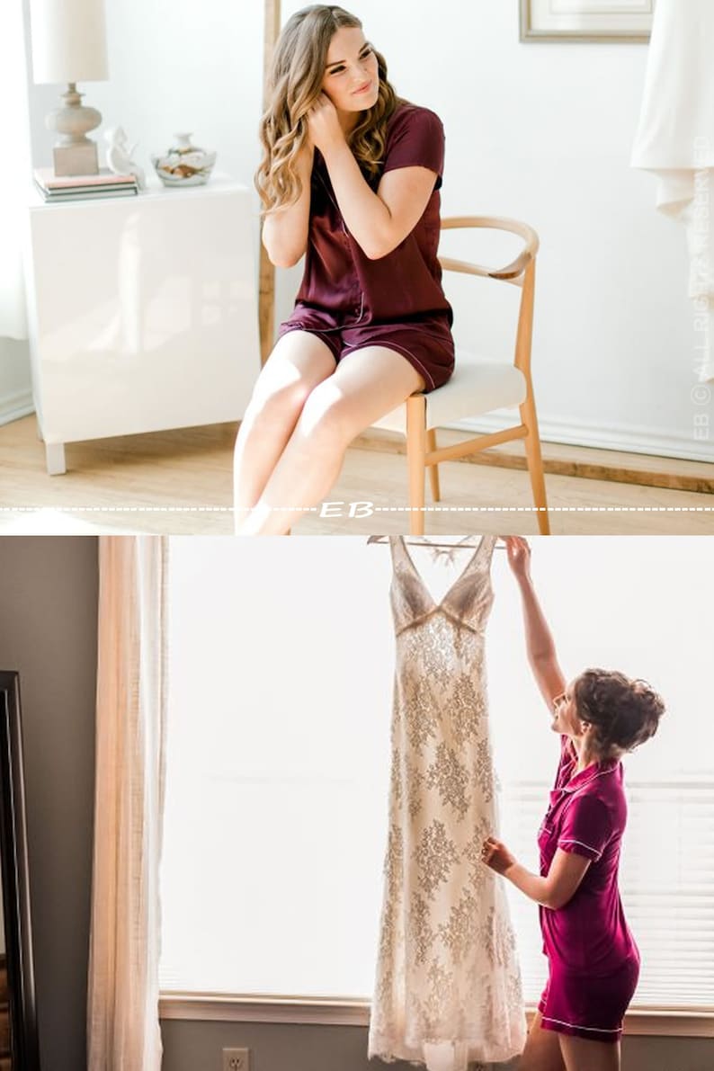 May include: Two people in matching burgundy pajamas getting ready for a wedding. One person is sitting on a chair and putting on earrings. The other person is holding up a white lace wedding dress.