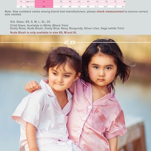 May include: Two girls wearing pink satin pajamas. The girl on the left is wearing a white pajama set and the girl on the right is wearing a pink pajama set. The size chart for the pajamas is shown in the background.
