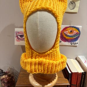 May include: A vibrant yellow knitted hat with cat ears. The hat, made from chunky yarn, has a ribbed brim and pointed ears. It's displayed on a mannequin head. The background includes artwork and books.