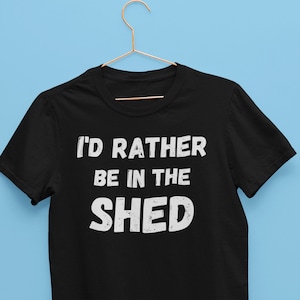 May include: Black t-shirt with the white text "I'D RATHER BE IN THE SHED". The shirt is hanging on a gold hanger against a light blue background. The text is in a bold, sans-serif font.