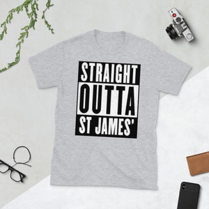 May include: A light gray t-shirt with the text "STRAIGHT OUTTA ST JAMES'" in a black and white rectangular design. The shirt is short-sleeved and has a crew neck. A camera, wallet, glasses, and phone are also visible.