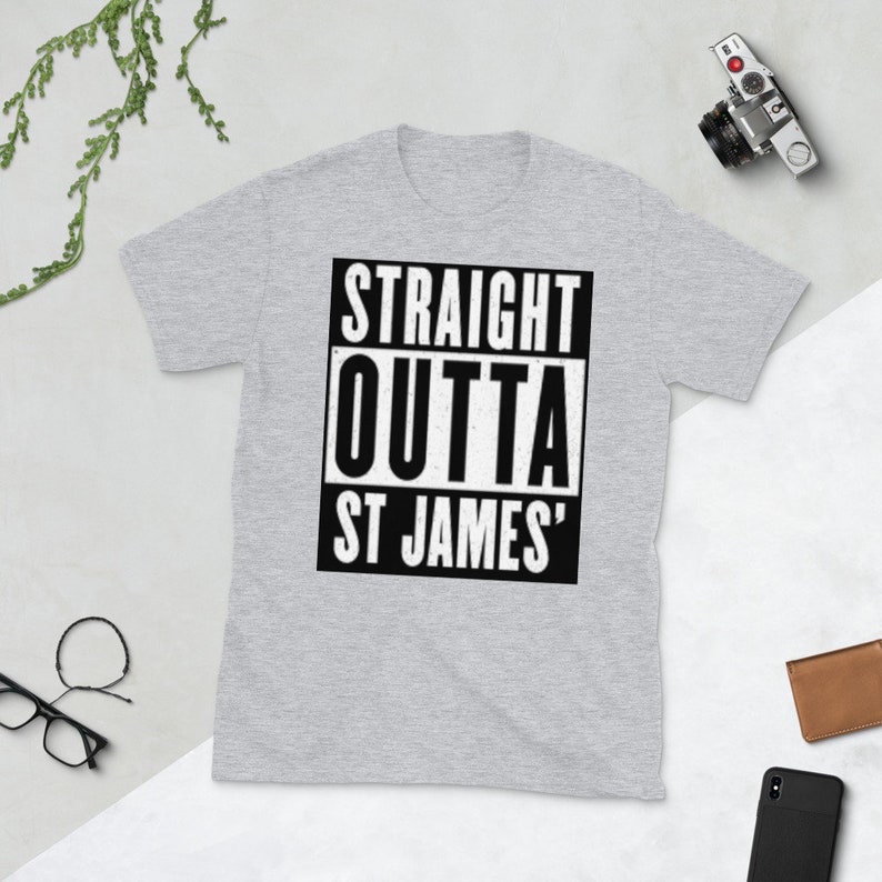 May include: A light gray t-shirt with the text "STRAIGHT OUTTA ST JAMES'" in a black and white rectangular design. The shirt is short-sleeved and has a crew neck. A camera, wallet, glasses, and phone are also visible.
