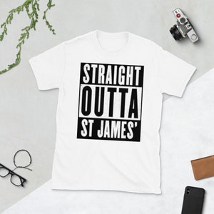 May include: White t-shirt with the text "STRAIGHT OUTTA ST JAMES'" in a black and white design. The text is in a bold, blocky font. The shirt is short-sleeved and appears to be made of a soft material. A camera, glasses, and wallet are in the background.