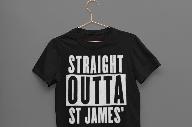 May include: Black t-shirt with the text "STRAIGHT OUTTA ST JAMES'" in a bold, white, distressed font. The shirt is hanging on a gold-colored hanger against a gray background. The t-shirt is a classic crew neck style.