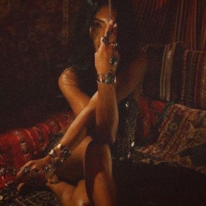 May include: A person wearing multiple silver rings and bracelets with green stones. The person is sitting on a red and patterned cushion. The image has a warm, vintage aesthetic.