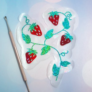 Pu&ograve; includere: Un adesivo trasparente e iridescente con un design a vite di fragole con fragole rosse e foglie verdi glitterate. Uno strumento argentato &egrave; posizionato a sinistra, suggerendo un progetto fai-da-te.