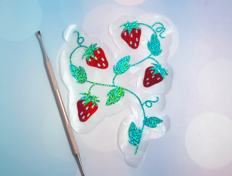Pu&ograve; includere: Un adesivo trasparente e iridescente con un design a vite di fragole con fragole rosse e foglie verdi glitterate. Uno strumento argentato &egrave; posizionato a sinistra, suggerendo un progetto fai-da-te.