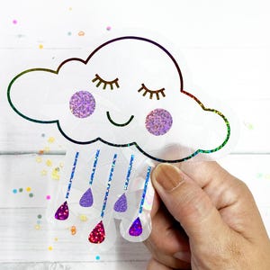 May include: A rainbow iridescent vinyl sticker of a smiling cloud with closed eyes and raindrops. The cloud has glitter accents on the cheeks.