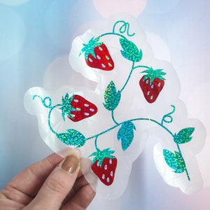 Pu&ograve; includere: Un adesivo trasparente fustellato con un viticcio di fragole con quattro fragole rosse e foglie verdi. Le foglie e i viticci hanno una finitura glitterata, verde acqua e le fragole hanno dettagli di semi bianchi.