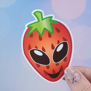 Può includere: Un adesivo a forma di fragola rossa con un gambo e foglie verdi. La fragola ha un viso alieno da cartone animato con due grandi occhi neri e una bocca sorridente.