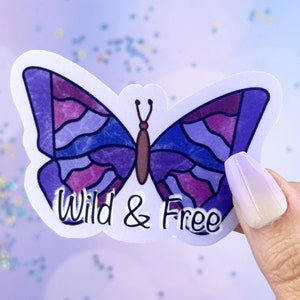 Può includere: Un adesivo a forma di farfalla viola e blu con un design a vetri colorati. L'adesivo ha il testo "Wild & Free" scritto sotto la farfalla.