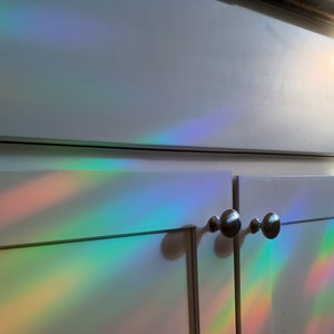 Pu&ograve; includere: Ante di armadi bianche con pomelli argentati illuminate da uno spettro di luce arcobaleno. La luce crea bande colorate di rosso, arancione, giallo, verde, blu e viola sulla superficie dell'armadio.