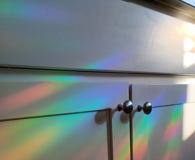 Pu&ograve; includere: Ante di armadi bianche con pomelli argentati illuminate da uno spettro di luce arcobaleno. La luce crea bande colorate di rosso, arancione, giallo, verde, blu e viola sulla superficie dell'armadio.