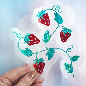 Pu&ograve; includere: Un adesivo trasparente con un design a vite di fragole. Le fragole sono rosse con semi bianchi, e le foglie e gli steli sono glitterati color teal. L'adesivo ha un bordo smerlato.