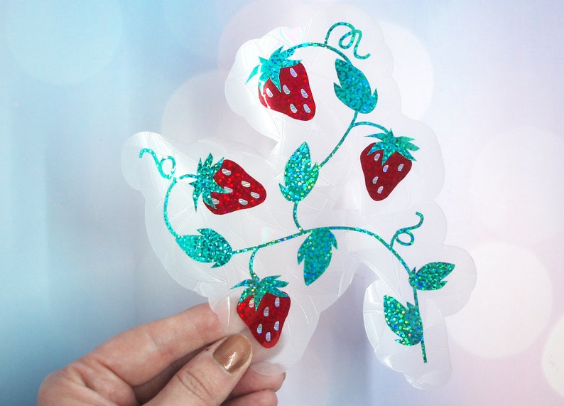 Pu&ograve; includere: Un adesivo trasparente con un design a vite di fragole. Le fragole sono rosse con semi bianchi, e le foglie e gli steli sono glitterati color teal. L'adesivo ha un bordo smerlato.