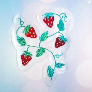Pu&ograve; includere: Un adesivo trasparente con un design a vite di fragole. Le fragole sono rosse con riflessi bianchi, e le foglie e i viticci sono verdi con accenti glitterati. L'adesivo ha un effetto olografico.
