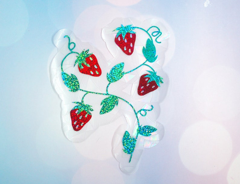 Pu&ograve; includere: Un adesivo trasparente con un design a vite di fragole. Le fragole sono rosse con riflessi bianchi, e le foglie e i viticci sono verdi con accenti glitterati. L'adesivo ha un effetto olografico.