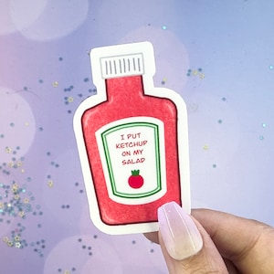 Può includere: Un adesivo a forma di bottiglia di ketchup rossa con un'etichetta bianca e testo verde che dice "Metto il ketchup sulla mia insalata". Un pomodoro rosso è illustrato sull'etichetta.