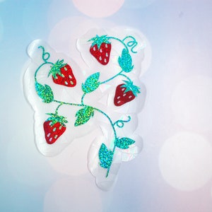 Pu&ograve; includere: Un adesivo trasparente con un design a vite di fragole. Le fragole sono rosse con semi bianchi, e le foglie e i viticci sono verdi con glitter. L'adesivo ha un contorno bianco irregolare.