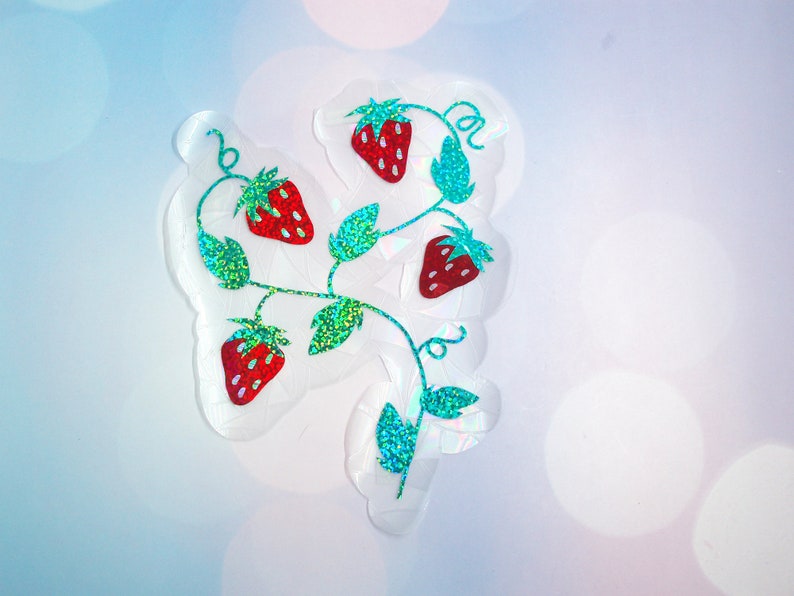 Pu&ograve; includere: Un adesivo trasparente con un design a vite di fragole. Le fragole sono rosse con semi bianchi, e le foglie e i viticci sono verdi con glitter. L'adesivo ha un contorno bianco irregolare.