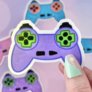 May include: A collection of colorful video game controller stickers. The central sticker is purple with green and black buttons. Other controllers are blue, pink, and purple. The stickers have a white border and are on a soft purple background.