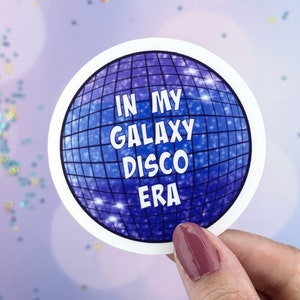 Può includere: Un adesivo rotondo con un design a palla da discoteca blu e il testo "In My Galaxy Disco Era".