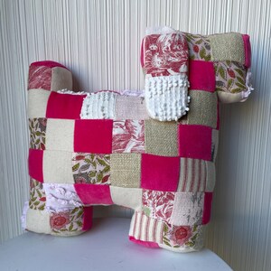 May include: A patchwork dog pillow with pink, white, and beige fabric squares. The pillow has a white crocheted ear and a floral pattern on the fabric.