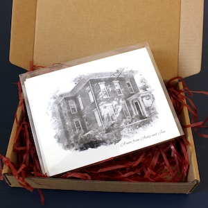 May include: A brown cardboard box with red shredded paper inside. The box is open and contains a white card with a black and white drawing of a house. The card has the text "A note from Aunt and Jim" printed on it.