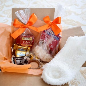 May include: A gift box containing tea, chocolates, and white fluffy socks. The box is lined with orange tissue paper and tied with orange bows. The box includes a Hershey's chocolate bar and a candle.