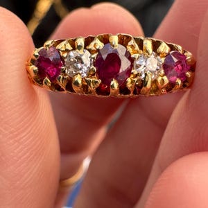 May include: A gold ring featuring a five-stone setting with three deep red rubies and two clear white diamonds. The stones are arranged in a row, held by gold prongs within a textured, ornate gold band that spans the width of the finger.