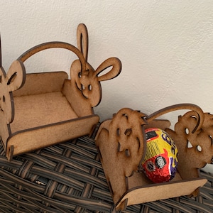 May include: Two wooden bunny-shaped Easter baskets. One basket is empty and the other holds a chocolate egg wrapped in gold foil.