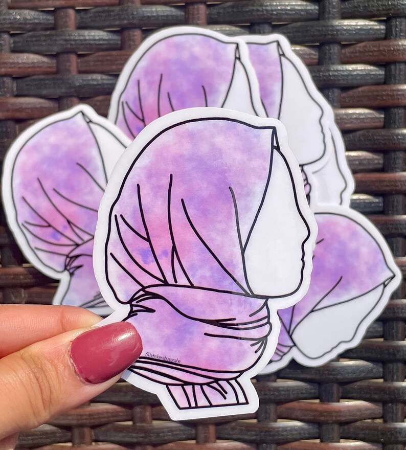 May include: Three purple and pink watercolour stickers of a woman wearing a headscarf. The stickers are on a woven brown background.