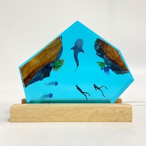 Könnte beinhalten: Dekorative Harzlampe mit einer blauen Ozeanszene. Die Lampe zeigt einen Walhai, Quallen und Taucher. Die Seiten der Lampe haben Holzakzente. Die Lampe steht auf einem rechteckigen Holzsockel.