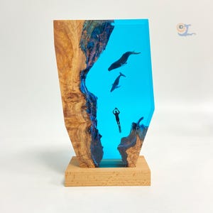 May include: A wooden sculpture with a blue resin inlay depicting a diver and two whales swimming in the ocean. The sculpture is on a wooden base.