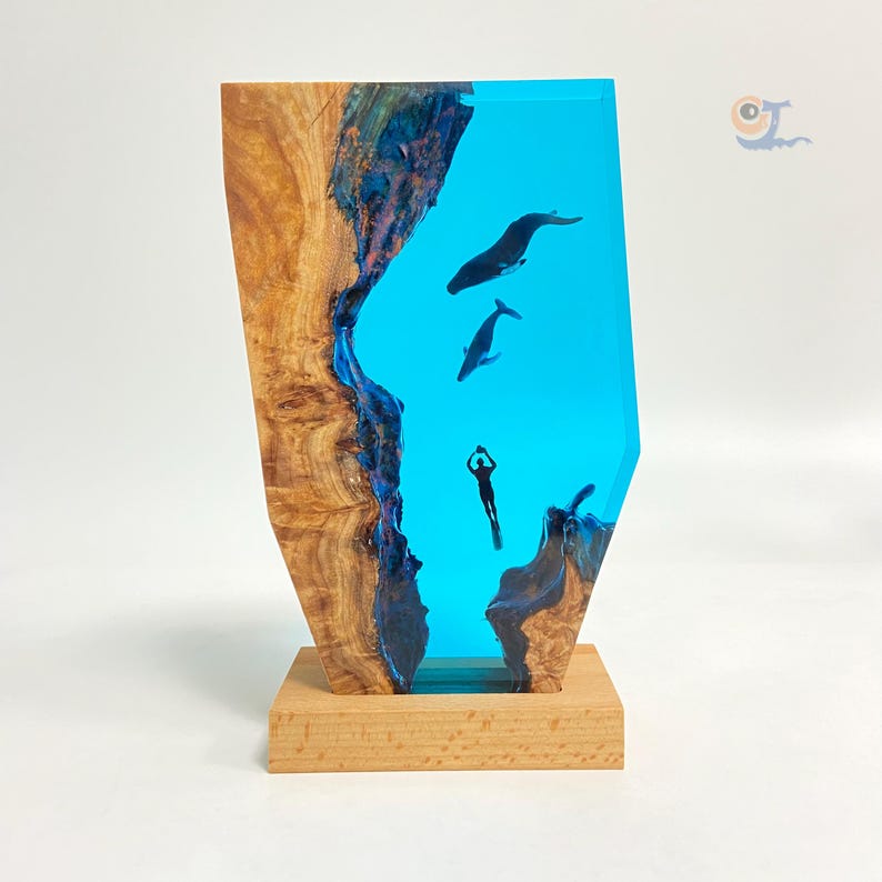 May include: A wooden sculpture with a blue resin inlay depicting a diver and two whales swimming in the ocean. The sculpture is on a wooden base.