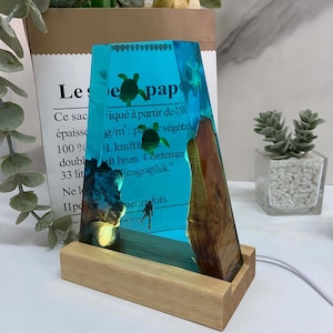 Op de afbeelding: Decoratieve harslamp met een turquoise interieur met zeeschildpadden en een duiker. De lamp heeft een houten voet en een bruine, houtachtige zijkant. De tekst "Le pap" is zichtbaar.