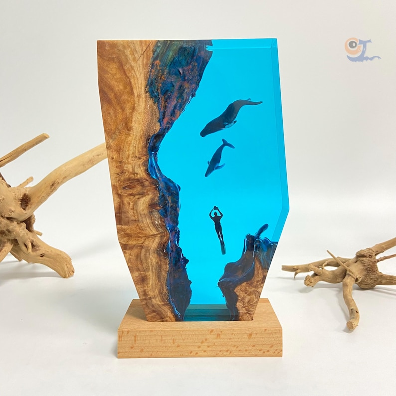 May include: A wooden sculpture with a blue resin inlay depicting a diver and whales. The sculpture is on a wooden base.