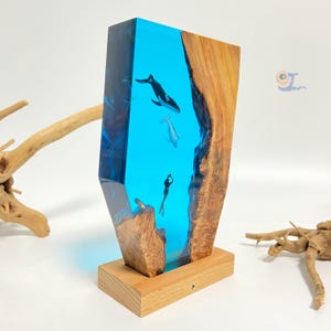 May include: A wooden sculpture with a blue resin insert depicting a whale, a dolphin, and a diver in a blue ocean. The sculpture is on a wooden base.