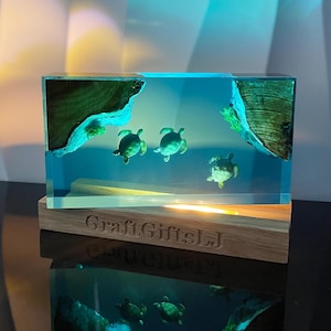 May include: A rectangular resin sculpture with a blue background and four green sea turtles. The sculpture is lit from below, casting a warm glow. The sculpture is on a wooden base with the text "Craft.Gifts.LJ" engraved on it.
