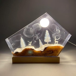 May include: A trapezoidal resin lamp featuring a natural wood base with a snowy landscape, white miniature deer, and pine trees. The clear resin upper section contains swirling white smoke patterns and a glowing circular light.