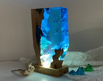 Könnte beinhalten: Eine blaue Harzlampe mit einem Holzsockel und einem Licht im Inneren. Das Harz ist klar und enthält ein Design von schwimmenden Fischen. Die Lampe steht auf einer weißen Oberfläche mit einer Muschel und grünen Glasobjekten.